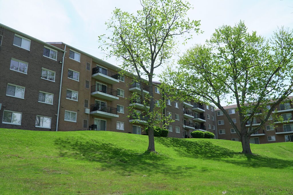 A multi-story brick apartment building in the North Hills set on a lush green hill with young green trees under a clear blue sky.