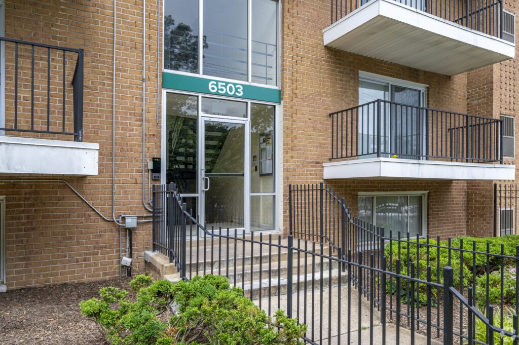Entrance to a brick apartment building with a green sign displaying the number 6503 above the glass door, flanked by metal railings, stairs, balconies, and trimmed bushes.
